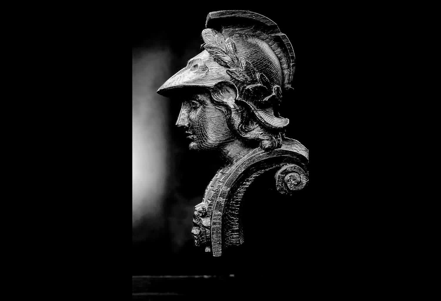 An ancient bronze bust in profile, lit from behind.